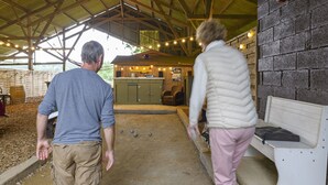 Un espacio al aire libre techado con suelo de tierra, bancos de madera y una zona de estar con cojines.