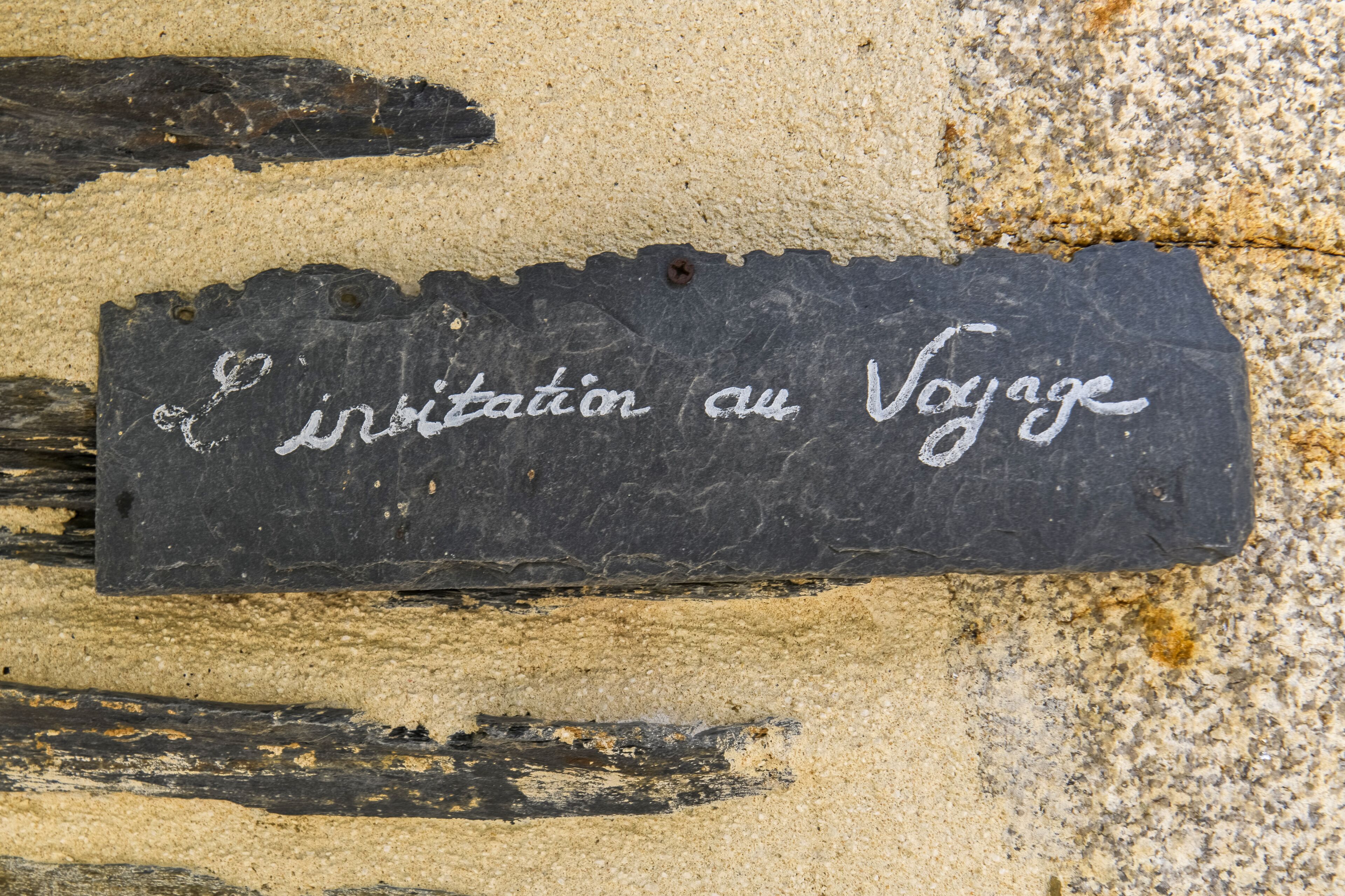 A black slate sign with white text reading "Invitation au Voyage" against a textured wall.