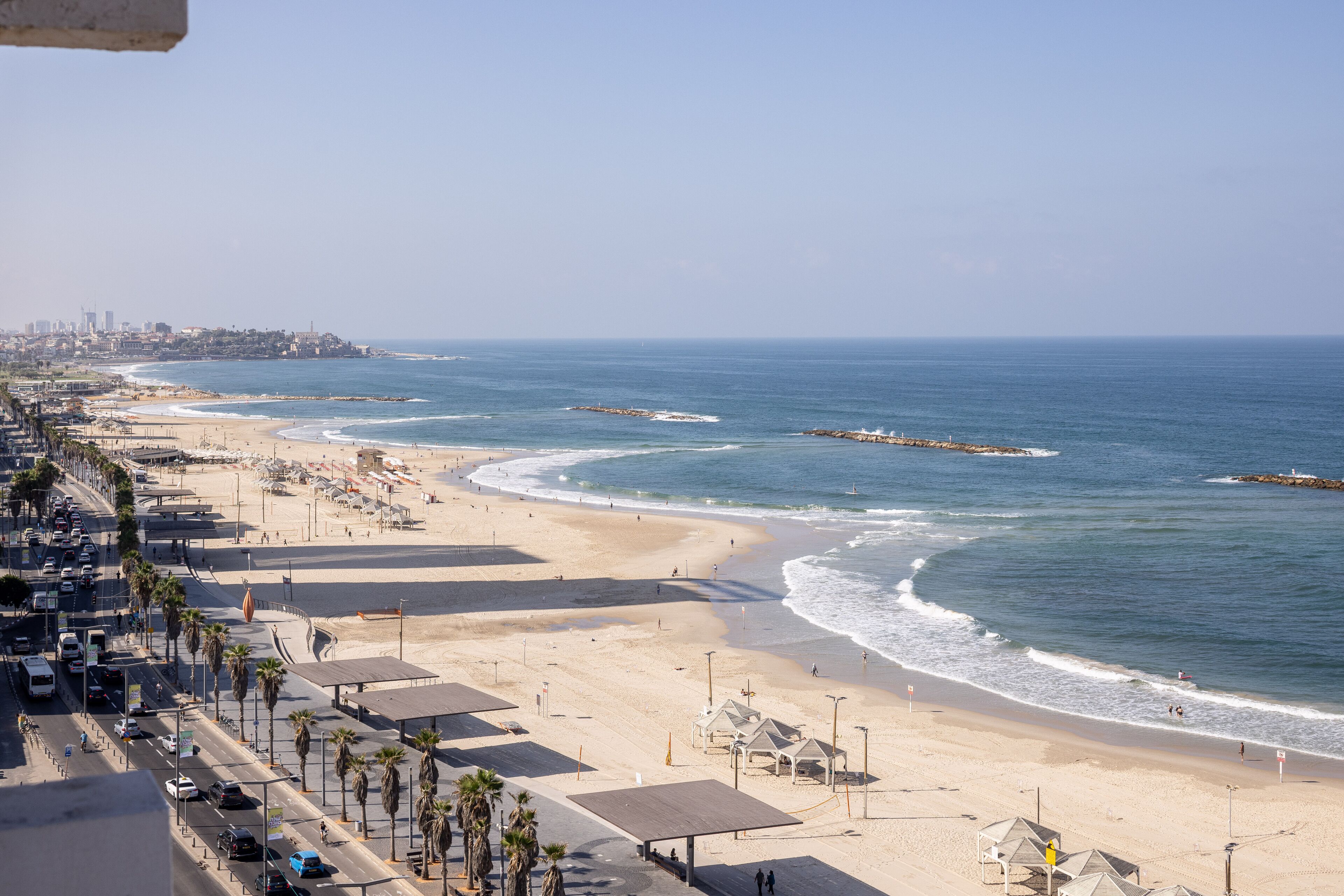 A coastal cityscape with a wide beach, palm-lined promenade, and a clear view of the ocean.