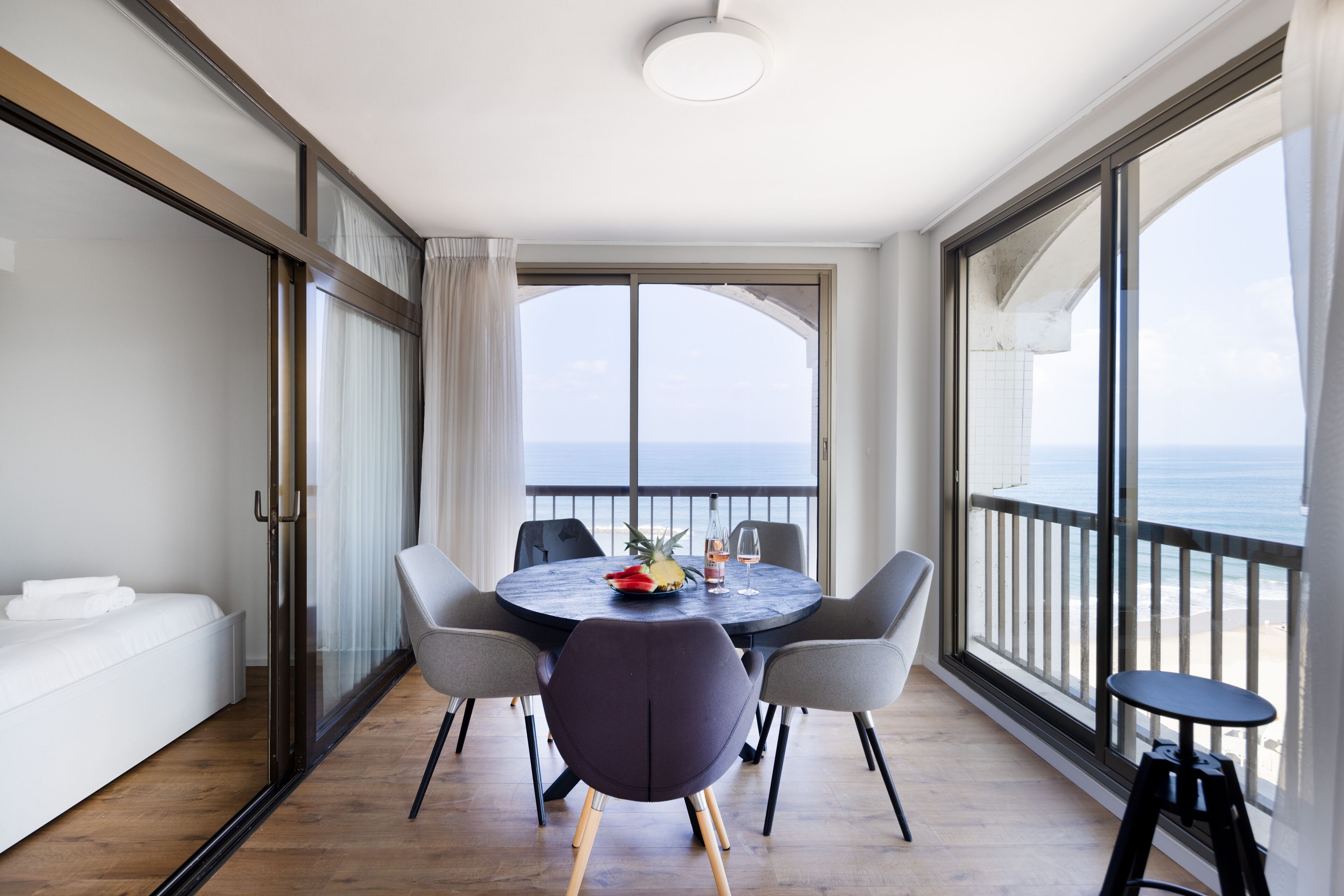 A modern room with a round table, chairs, a bed, and a large window offering a view of the sea.