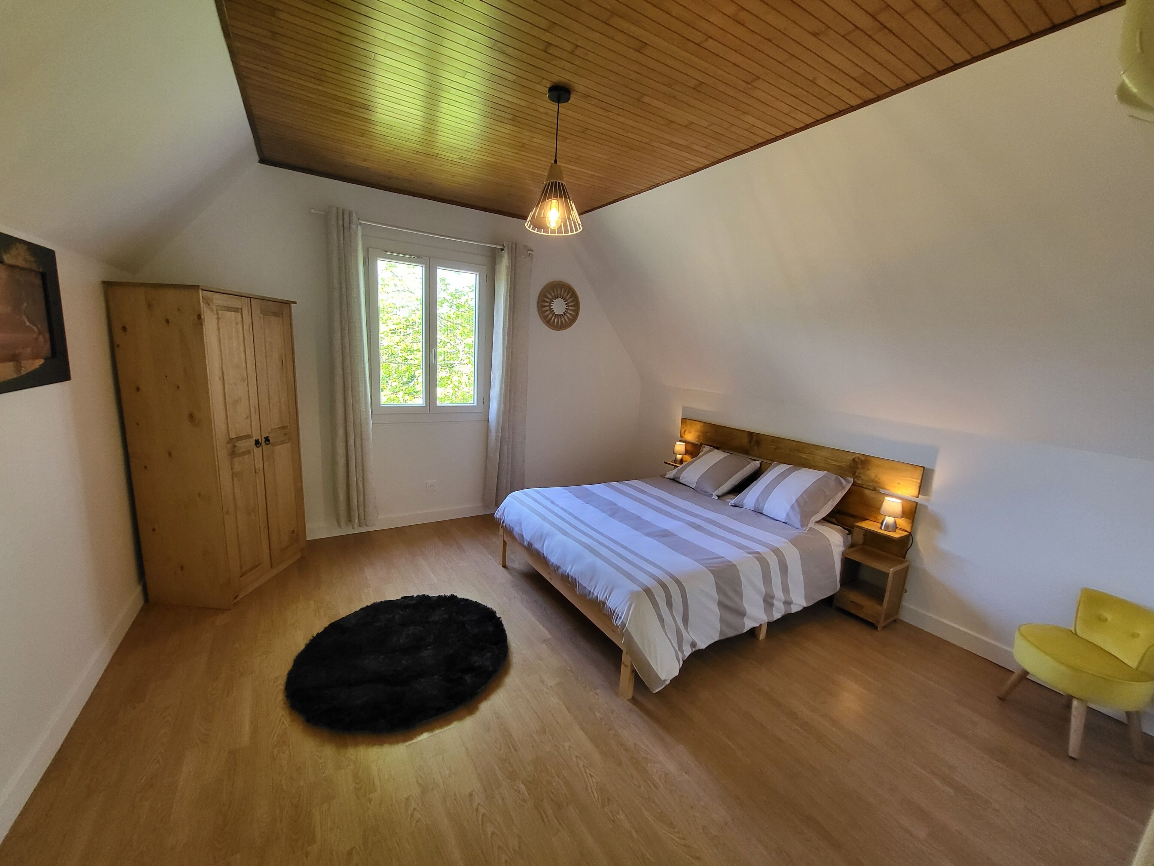 Camera da letto con soffitto in legno, un letto con biancheria a righe, un tappeto nero, un armadio in legno e una finestra con tende.