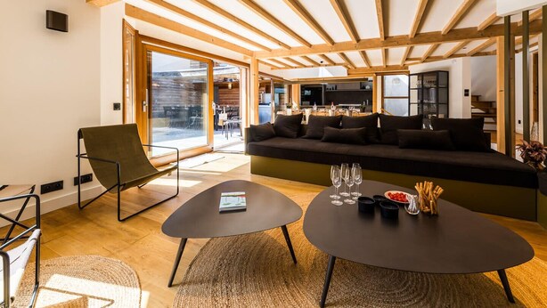 Una sala de estar moderna con techo de madera, un sofá, una mesa de centro y un comedor visible a través de puertas de vidrio.