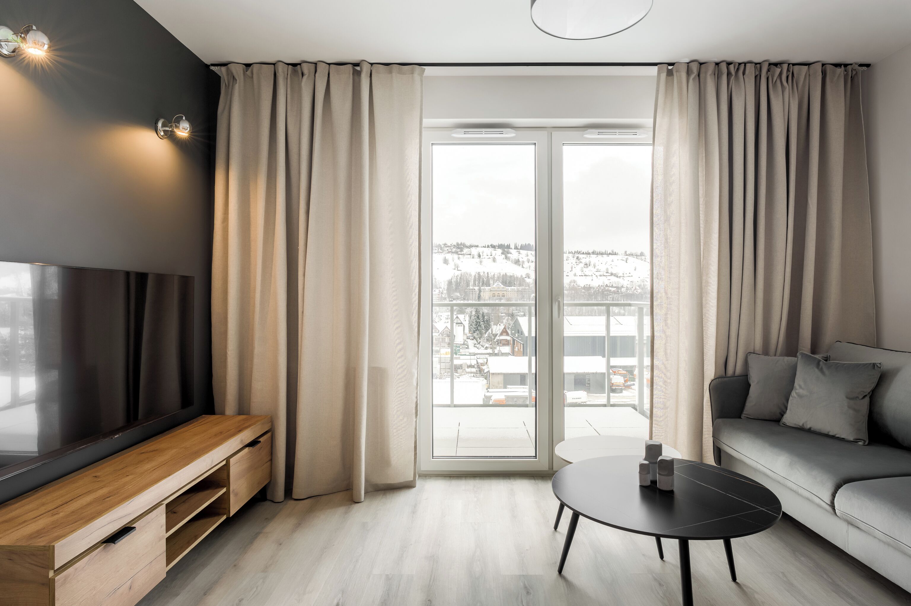 Uma sala de estar moderna com televisão, sofá e mesa de centro, grandes janelas com cortinas e vista para uma paisagem nevada.