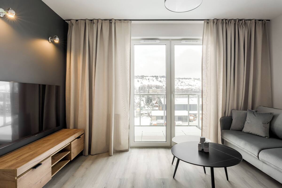A modern living room with a TV, sofa, and coffee table, large windows with curtains, and a view of a snowy landscape.
