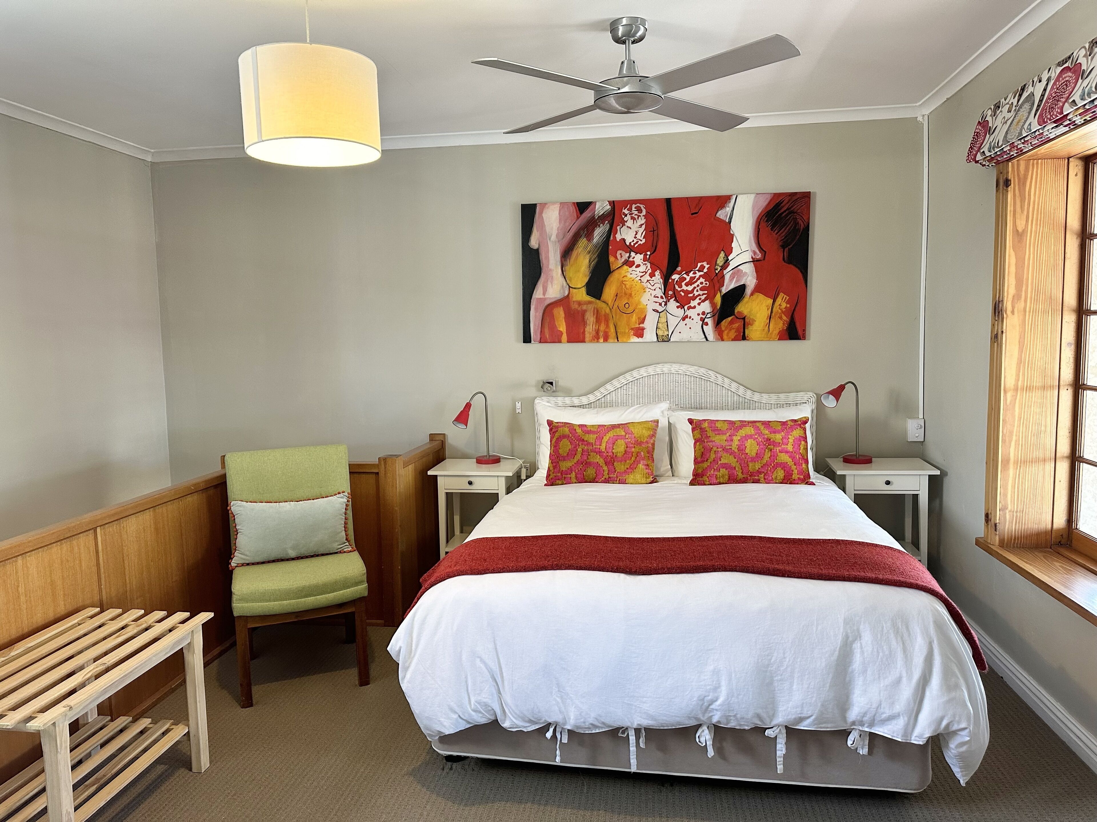 Un dormitorio con cama, una silla, mesita de noche con lámparas, ventilador de techo y un cuadro abstracto grande en la pared.