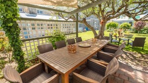 Een terras buiten met een houten tafel en rieten stoelen, omgeven door een wit lattenhek en groen.