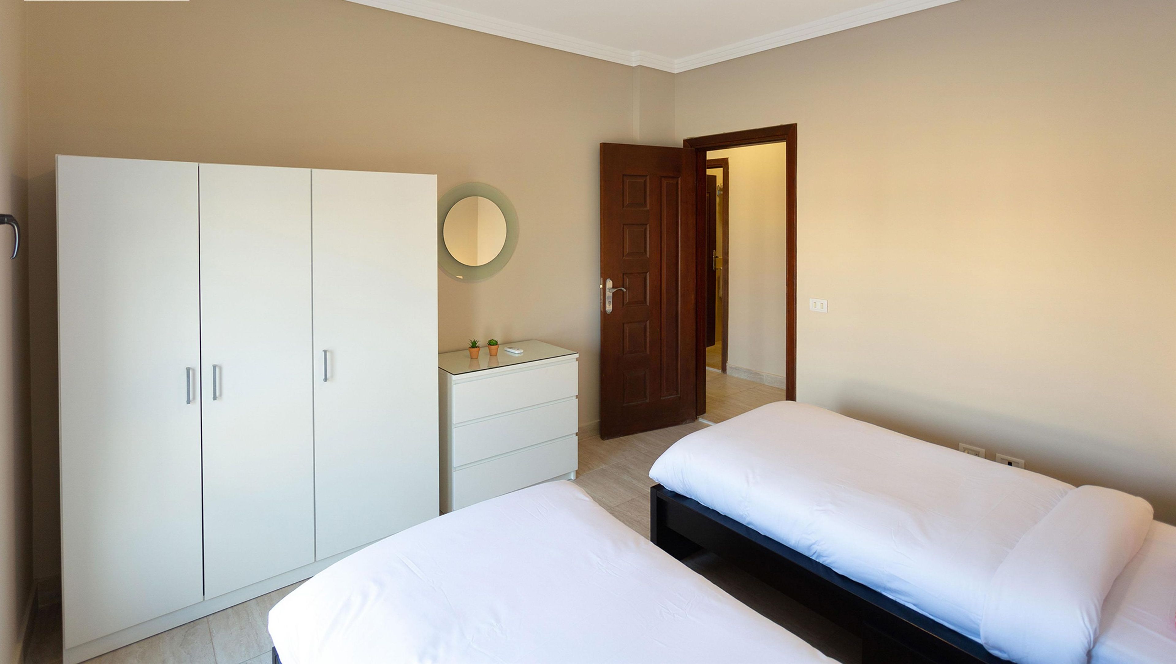 A bedroom with two beds, a wardrobe, a bedside table, and a mirror.