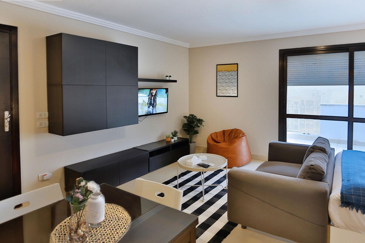 A modern living room with a flat-screen TV, a sofa, a coffee table, and a large window offering a city view.