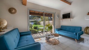 A living room with a blue sofa, a TV mounted on the wall, and a sliding glass door leading to a garden. - Préfailles - House 6P - 200m from the beaches (Préfailles)