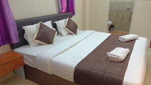 Una cama bien hecha con colcha marrón, ropa de cama blanca y dos almohadas. Un mesita de noche de madera con cajón está a la izquierda.