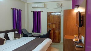 Habitación de hotel con cama, dos sillas, escritorio con televisor, unidad de aire acondicionado y cortinas moradas.