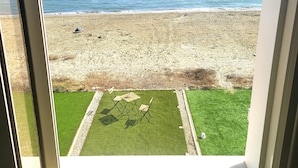 Vista de una playa a través de una ventana, que incluye una zona verde con una mesa y sillas.