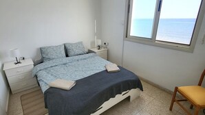 Un dormitorio con cama, mesita de noche, silla, ventana con vistas al mar y una lámpara en el techo.