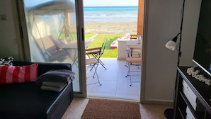 Habitación con puerta corrediza de vidrio que da a vista de la playa, sofá negro con almohada a rayas, mesa de madera con sillas y televisor sobre un mueble con un cartel de 'welcome'.