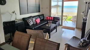 Una sala de estar moderna con un sofá de cuero negro, una mesa de centro y un comedor con sillas. Hay un ventanal con vista a la playa y al océano.