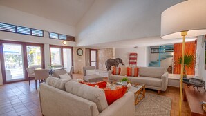 Een ruime woonkamer met een bank, fauteuils, een salontafel en een eethoek. Er is een groot raam, een klok aan de muur en een trap die naar een ander niveau leidt.