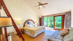 Een slaapkamer met een bed, nachtkastje, lamp, plafondventilator en uitzicht op een tuin via een glazen schuifdeur.