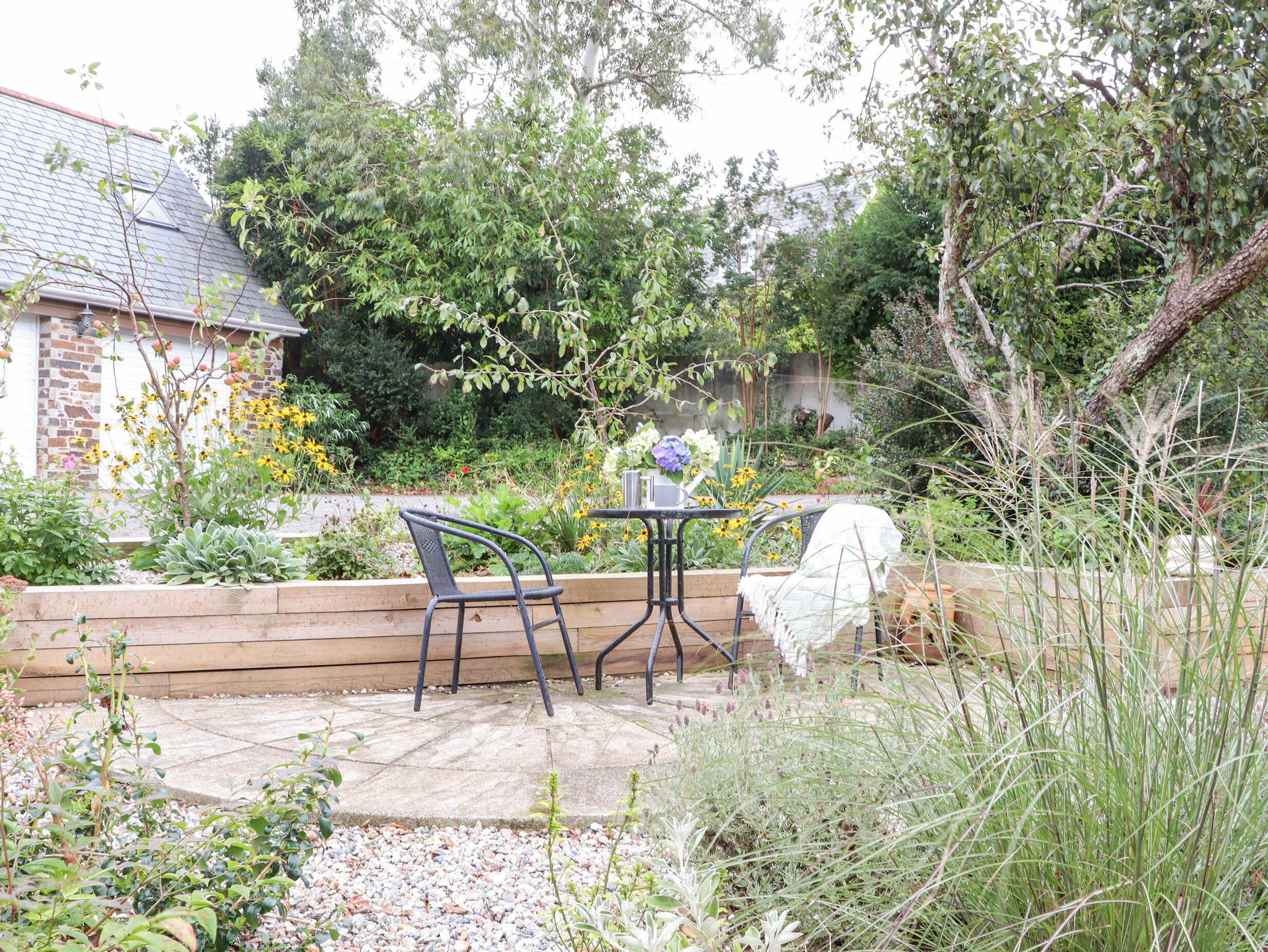 A patio with a table and chairs, surrounded by a garden with various plants and a brick wall in the background.