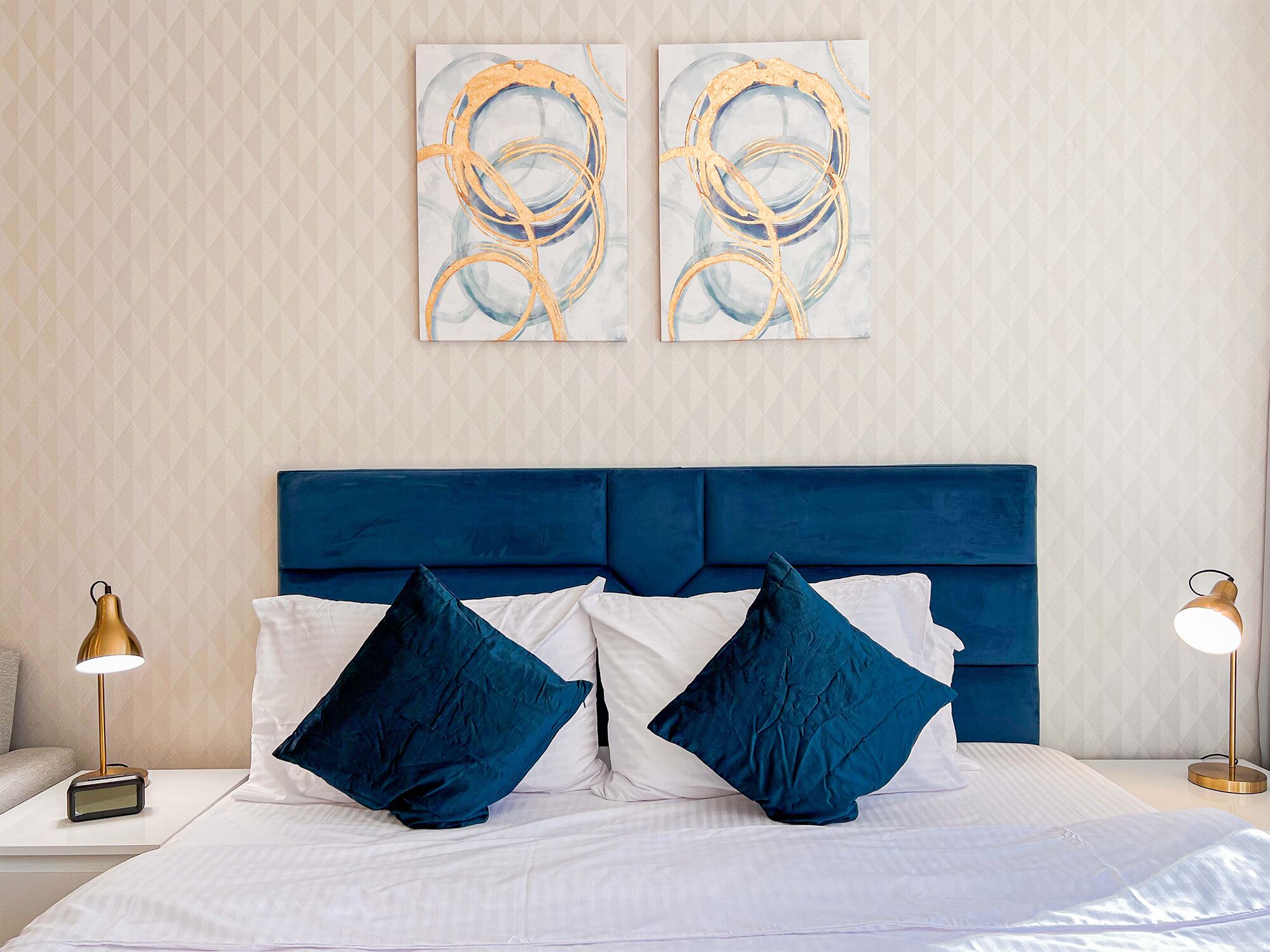 A hotel room with a large bed, two blue pillows, a white comforter, and two abstract paintings on the wall.