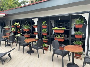 Un espace extérieur aménagé avec plusieurs tables et chaises, entouré de plantes en pot disposées sur des étagères.