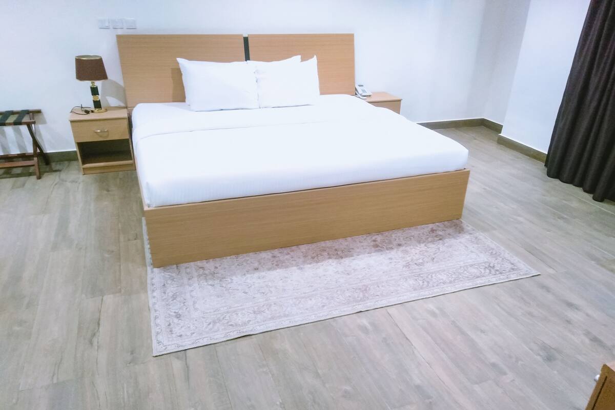 Un lit bien fait, avec une tête de lit en bois, une table de chevet avec une lampe et un tapis à motifs sur un plancher en bois.