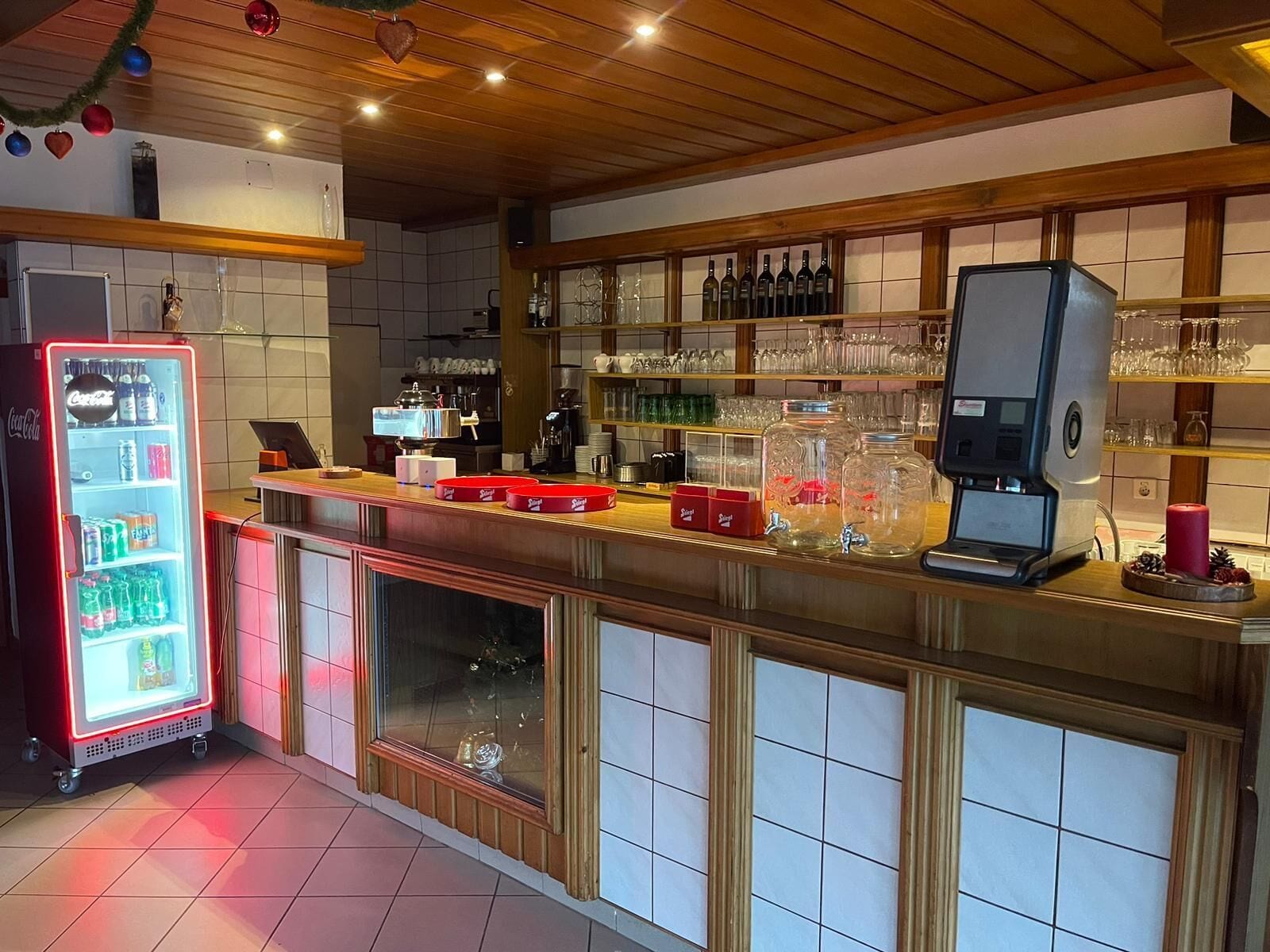 Un espace bar équipé d’un réfrigérateur Coca-Cola, d’une machine à café et d’étagères remplies de bouteilles et de verres.