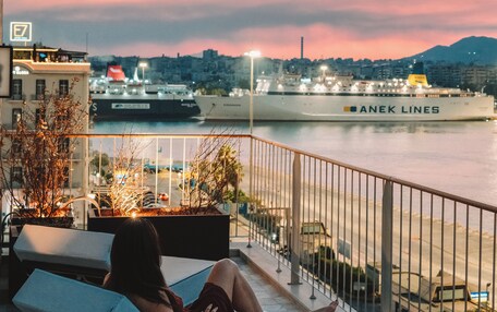 A balcony with a view of a waterfront, a person sitting on a lounge chair, and a large ship named 'ANEK LINES' in the background.