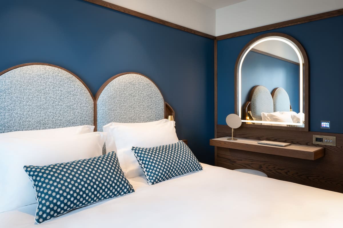 A bedroom with a large bed, two pillows, a nightstand, a mirror, and a wall-mounted control panel.