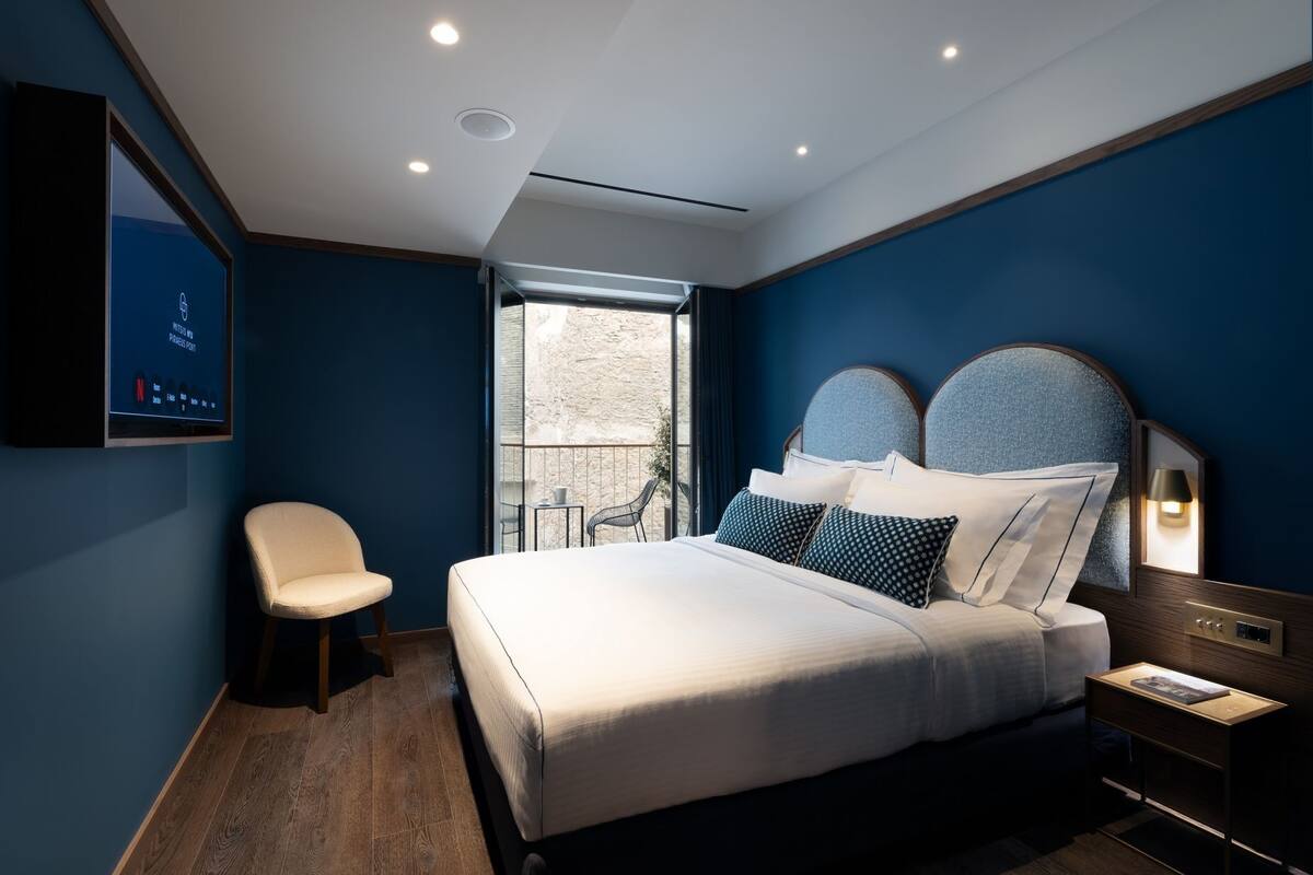 A modern hotel room with a large bed, a chair, a bedside table, and a TV mounted on the wall.