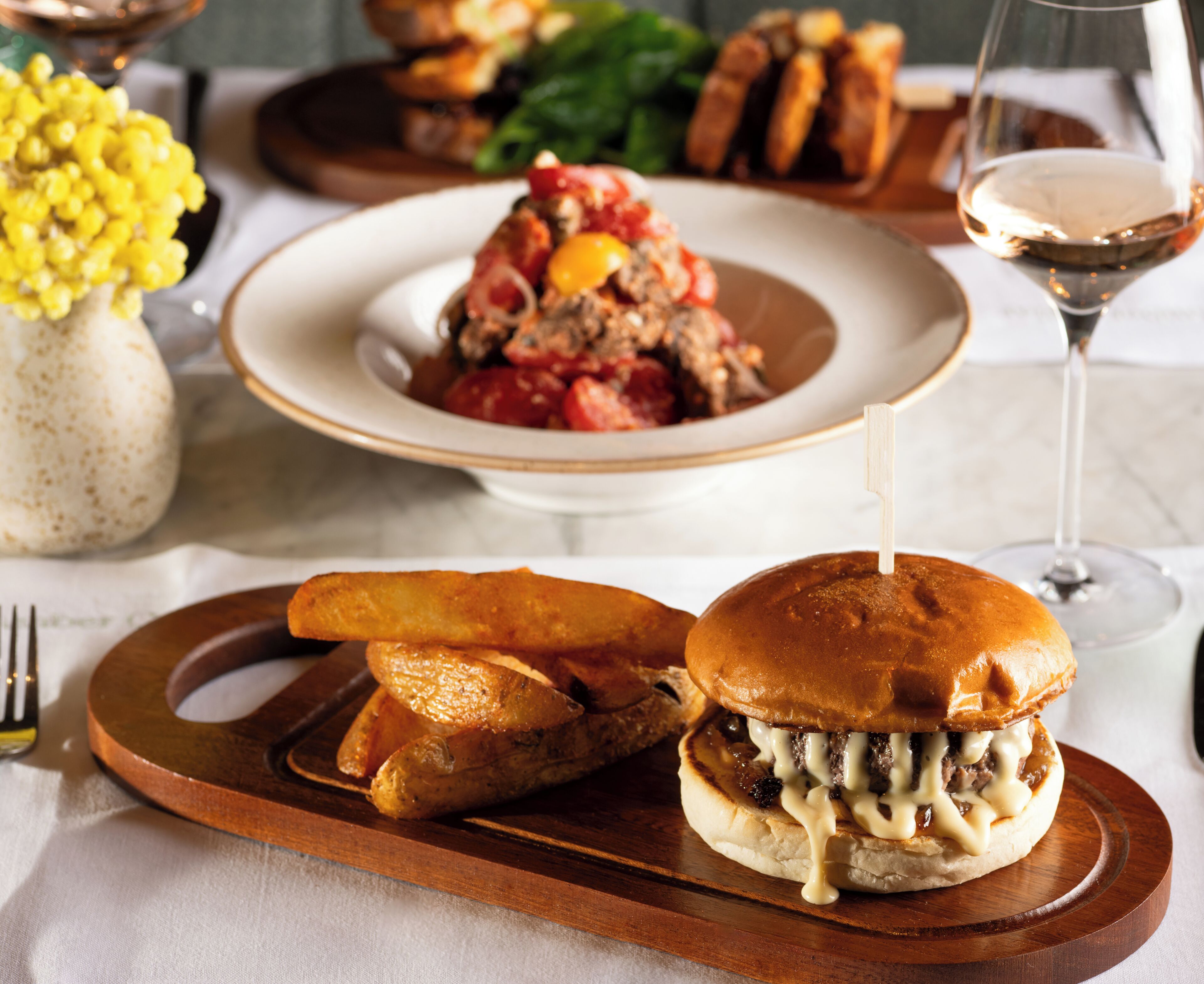 A meal with a burger and fries on a wooden board, a bowl of salad, and a glass of wine on a table.