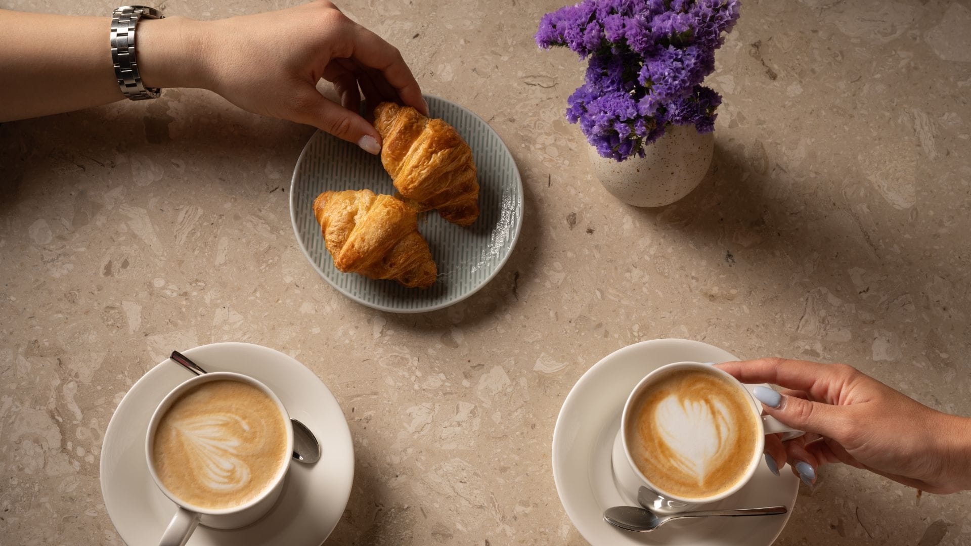 A table with croissants, two cups of coffee, and a vase of purple flowers.
