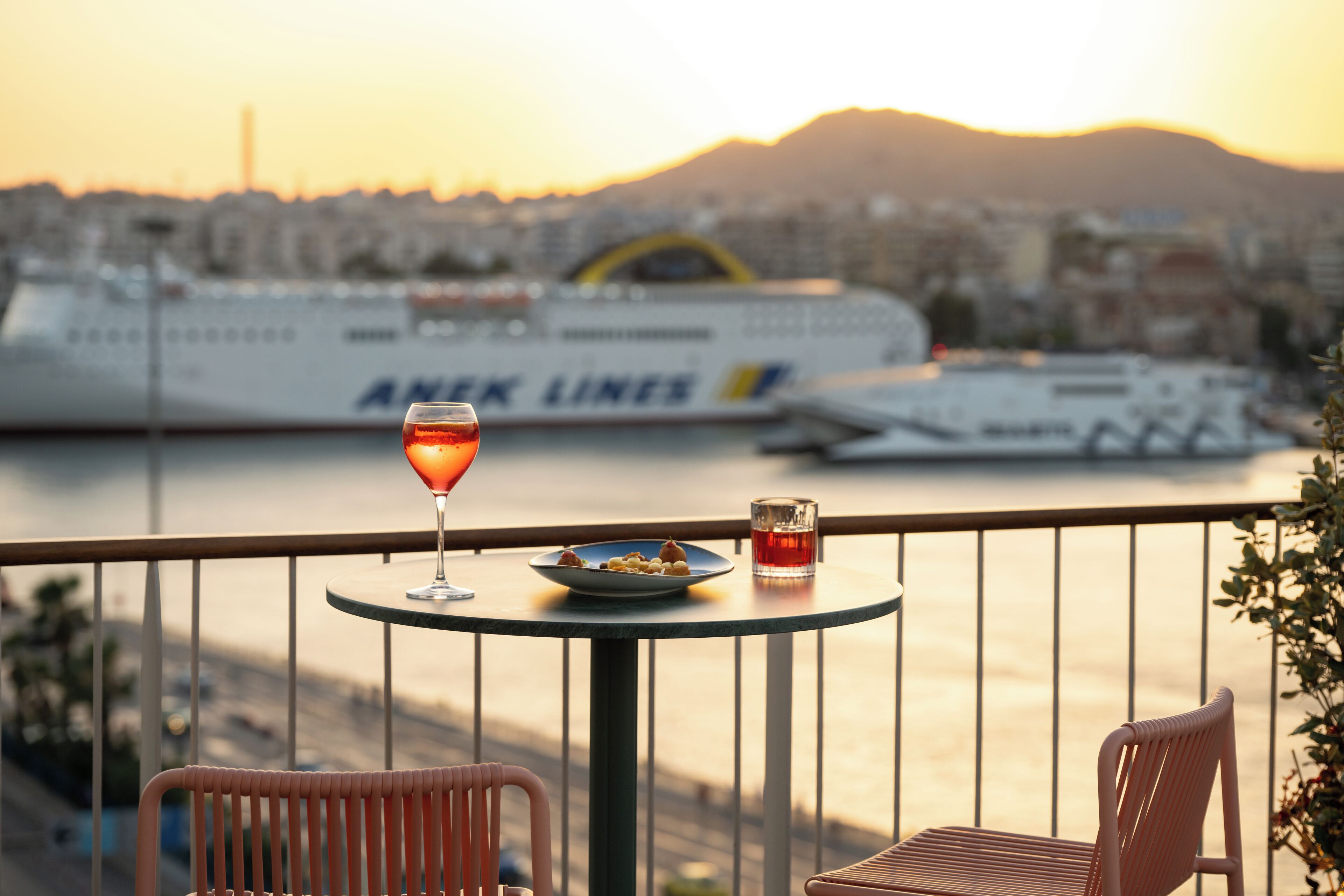 A balcony with a table, two chairs, a plate of food, a glass of drink, and a ship in the background.