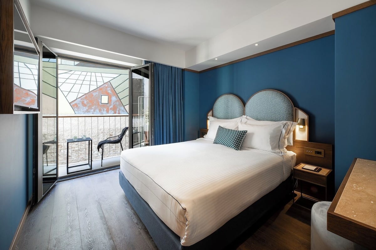 A modern hotel room with a large bed, a balcony with a chair, and a view of a building.