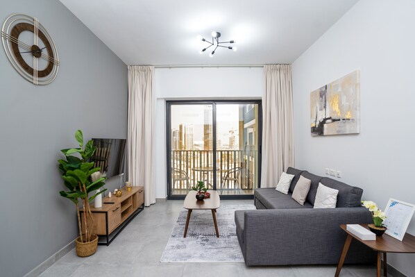 A modern living room with a grey sofa, a coffee table, a TV, and a balcony with a view of the city.