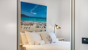 A bedroom with a bed, white bedding, a lamp, and a beach-themed wall art.