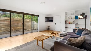 A modern living room with a large sliding glass door to a garden, a flat-screen TV, a sofa, a coffee table, and a bookshelf.