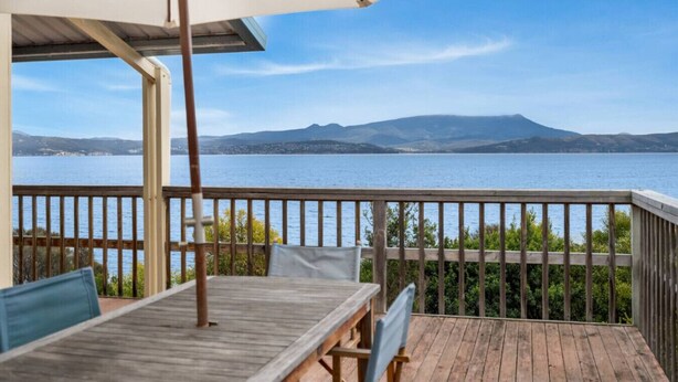 Una terraza de madera con una mesa y sillas, con vista a un cuerpo de agua y montañas al fondo.