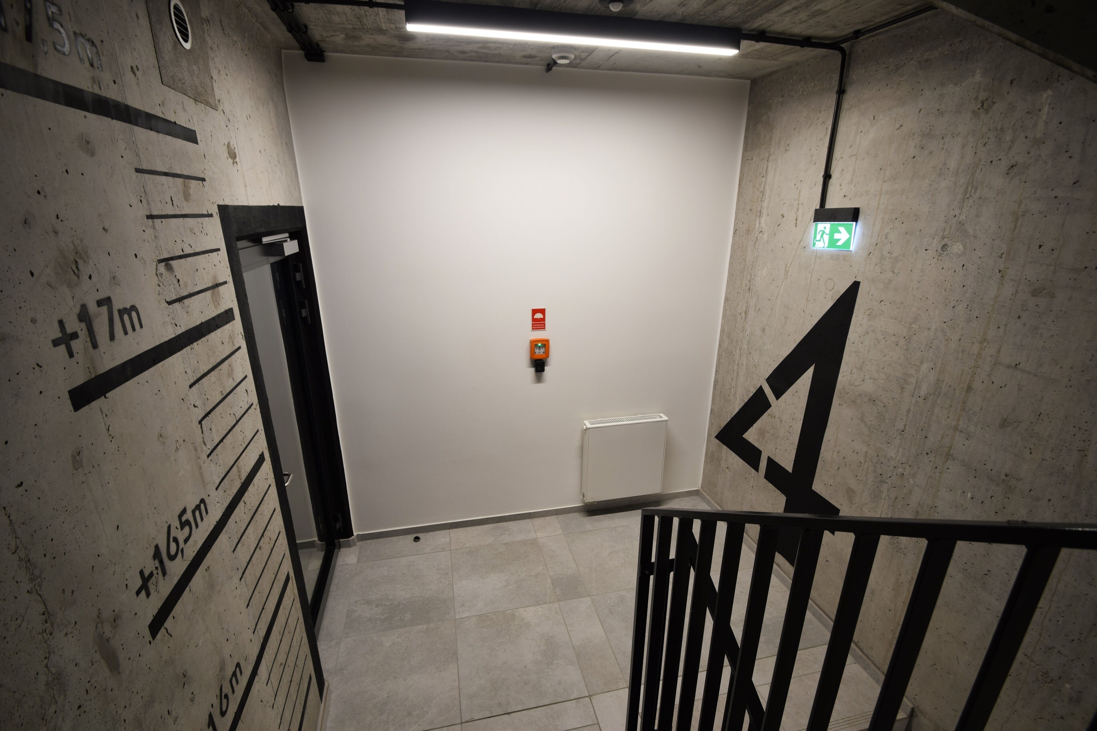 A stairwell with a handrail, a white door with an emergency exit sign, and a height measurement chart on the wall.