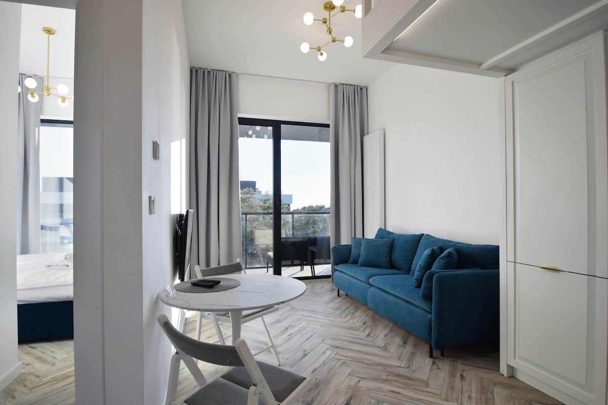 Uma sala de estar moderna com um sofá azul, uma mesa de jantar redonda com cadeiras e uma janela grande com cortinas.