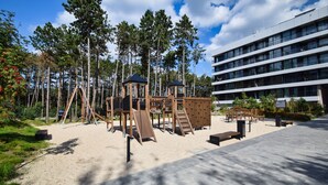 A playground with climbing structures and slides, surrounded by trees and a building in the background. - RentPlanet - Shellter (Trzebiatow)