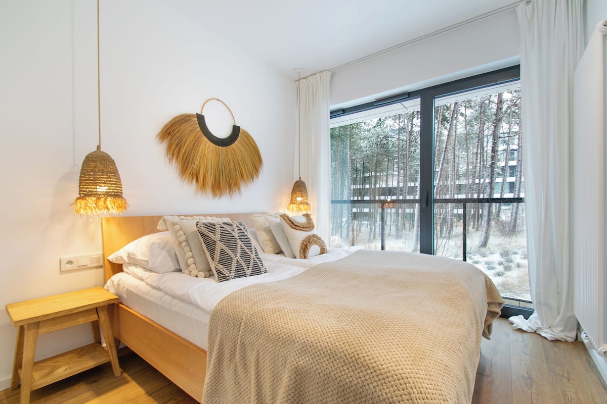 Quarto com uma cama grande, luminárias laterais, mesa de cabeceira de madeira e vista para uma paisagem nevada através de uma grande janela.
