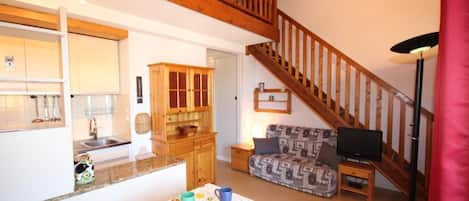 A cozy living room with a sofa, a TV, and a dining table with colorful cups. There's a wooden staircase leading upstairs.