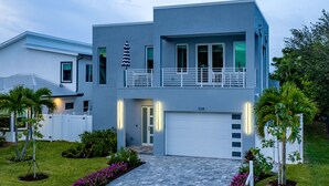 A modern two-story house with a garage, a balcony, and a well-maintained lawn with flowers and palm trees. - Elegant New Construction Naples Home w/ Heated Pool & Balcony (Naples)
