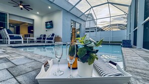 A poolside area with lounge chairs, a glass table with a bottle and glasses, and a large window overlooking the pool. - Elegant New Construction Naples Home w/ Heated Pool & Balcony (Naples)