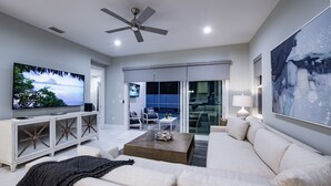 A modern living room with a flat-screen TV, a ceiling fan, a large painting, and a sliding glass door leading to a balcony. - Elegant New Construction Naples Home w/ Heated Pool & Balcony (Naples)