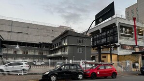 Un immeuble à plusieurs étages, avec une clôture blanche devant, une voiture noire et une voiture rouge garées dans la rue.
