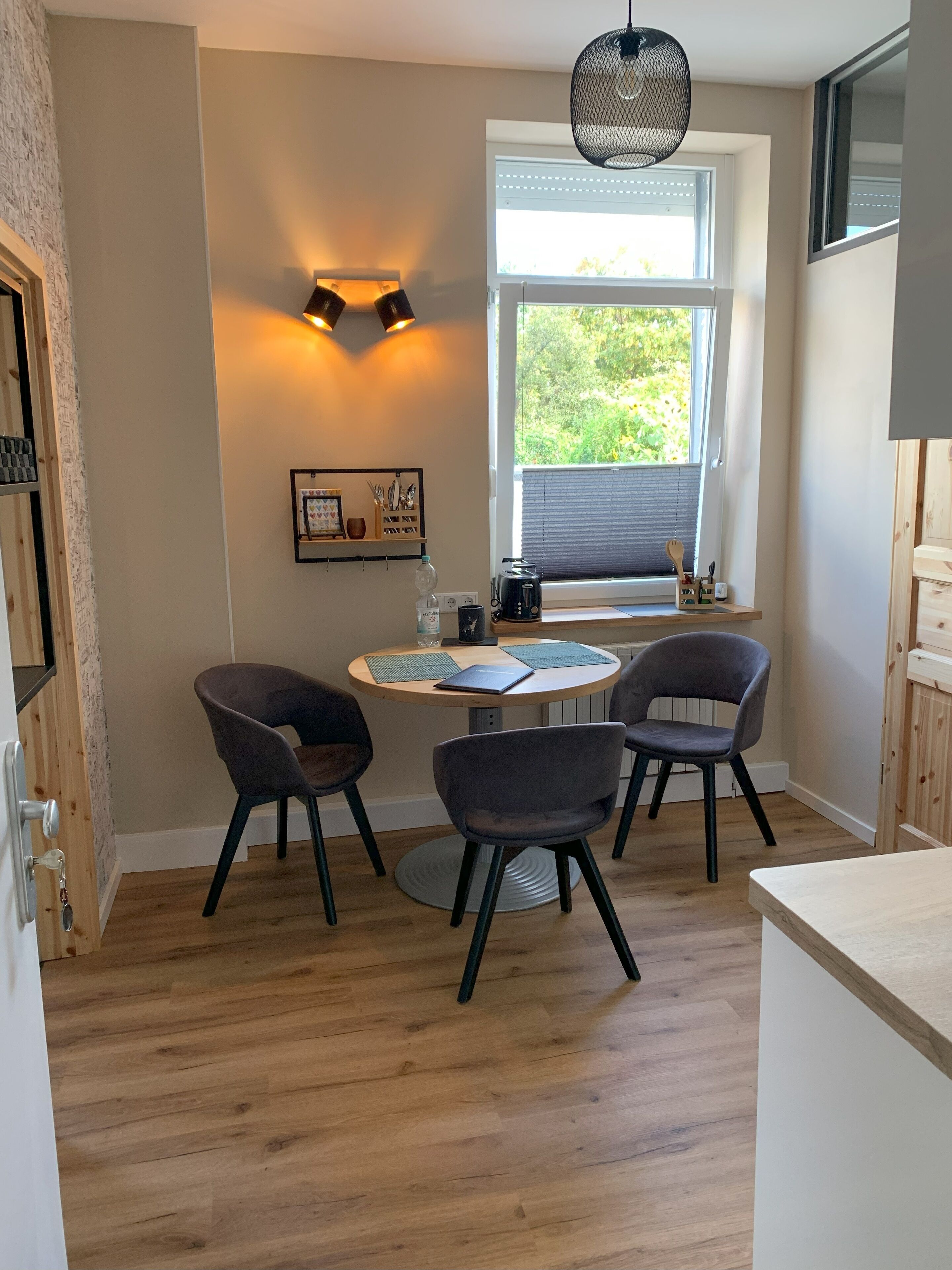 Une cuisine chaleureuse avec une table ronde, quatre chaises, un luminaire suspendu et une fenêtre avec des stores.
