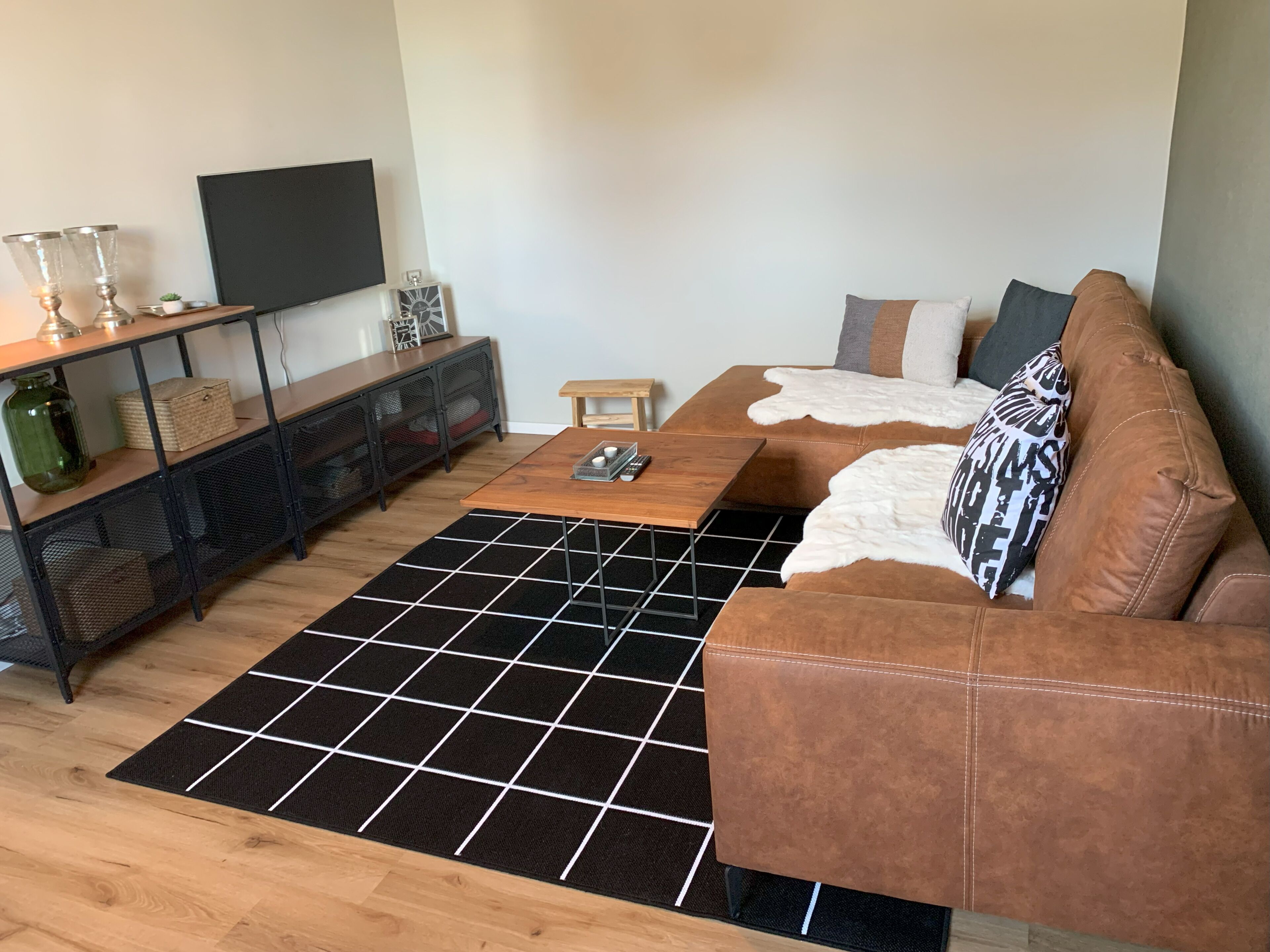 Un salon avec un canapé en cuir marron, une table basse en bois, un tapis à carreaux noirs et blancs, un téléviseur fixé au mur et une console en métal noir dotée d’étagères.