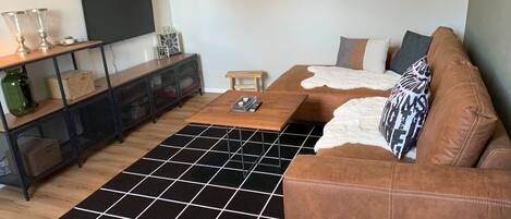 Un salon avec un canapé en cuir marron, une table basse en bois, un tapis à carreaux noirs et blancs, un téléviseur fixé au mur et une console en métal noir dotée d’étagères.