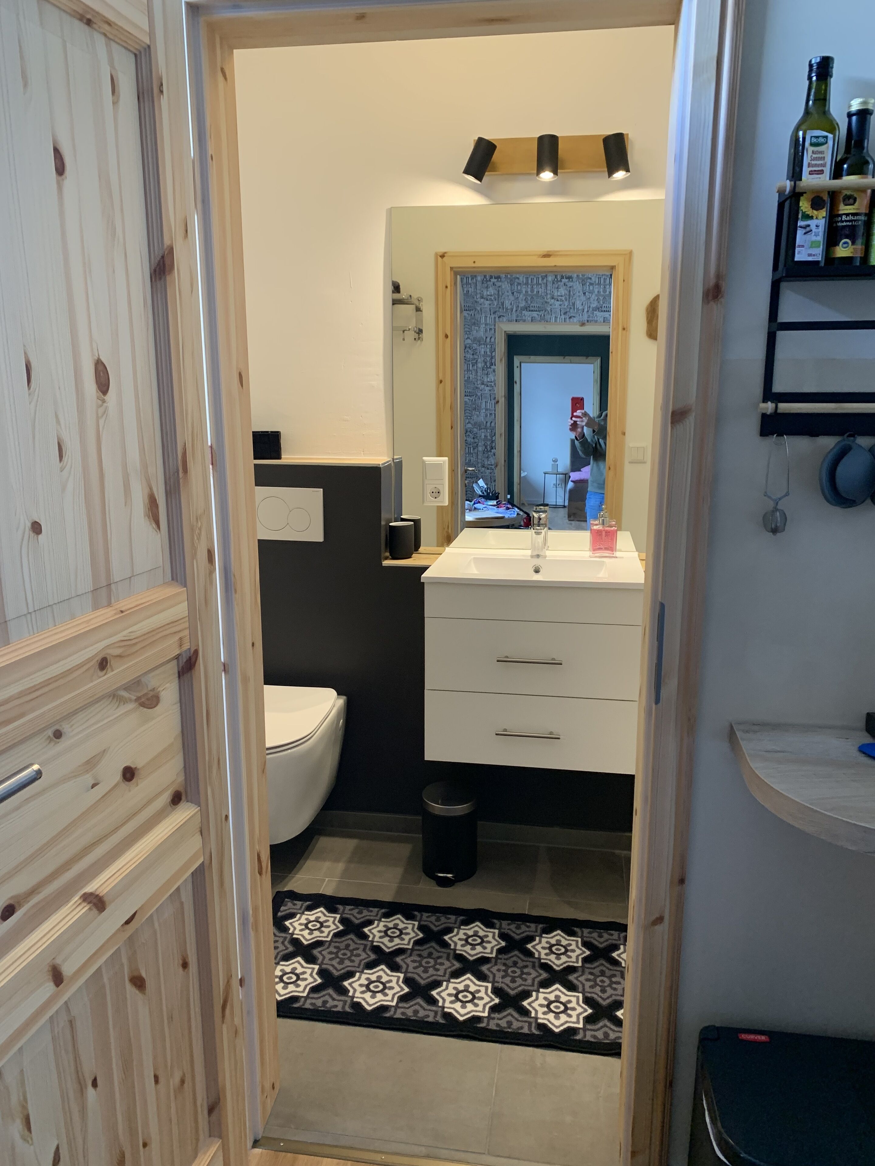 A bathroom with a white sink, a mirror, and a toilet. There is a black trash bin and a patterned rug on the floor.